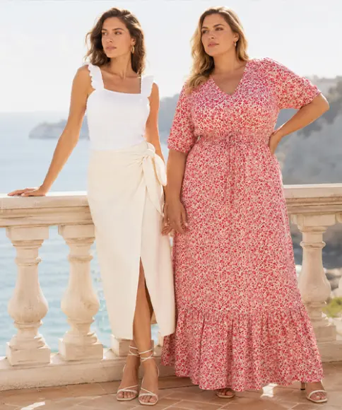 Twee vrouwen staan op een terras aan zee, één in een witte top met een crèmekleurige rok en de ander in een rode bloemen maxi-jurk, met kliffen op de achtergrond.